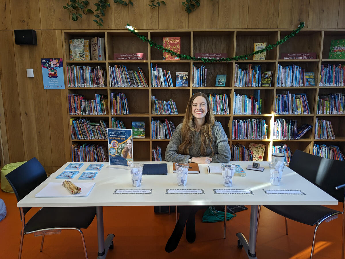 Fiona is sitting behind a table, smiling. A wall of bookshelves is visible behind her and a series of cups filled with paper are set out on the table in front of her
