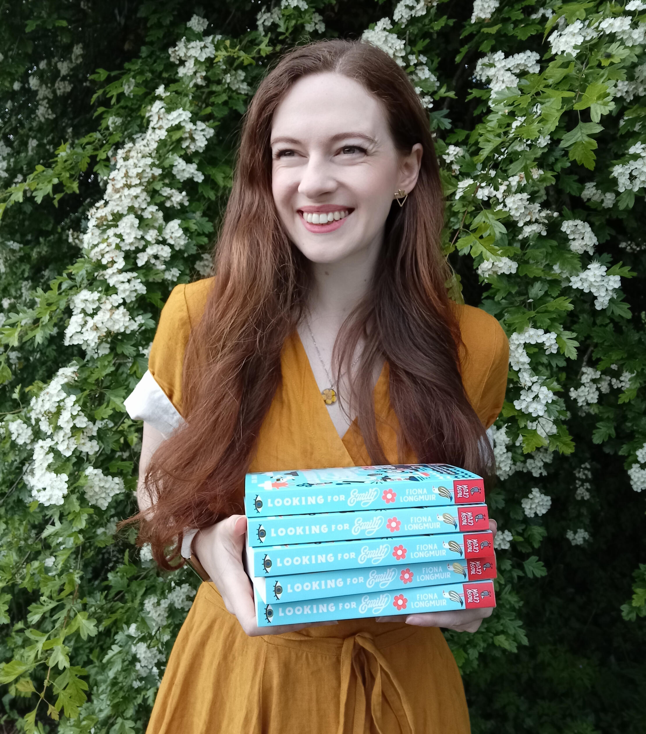 Fiona is looking to the side of the camera and laughing. She is holding a stack of Looking for Emily books and wearing a yellow linen dress. She is standing in front of a hawthorn tree covered in frothy white blossom.
