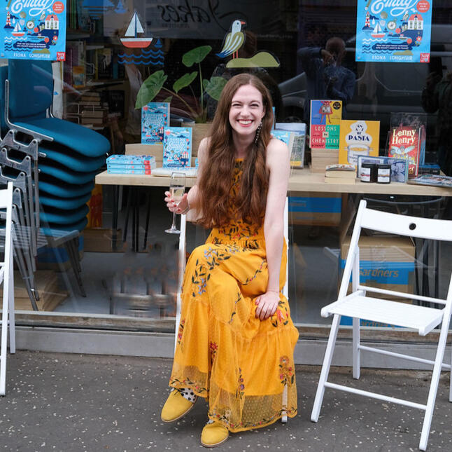 Fiona is sitting outside Mount Florida books wearing an embroidered yellow dress, holding a glass of champagne and beaming at the camera. The bookshop window is covered in Looking for Emily decals and posters. Copies of Looking for Emily are visible.