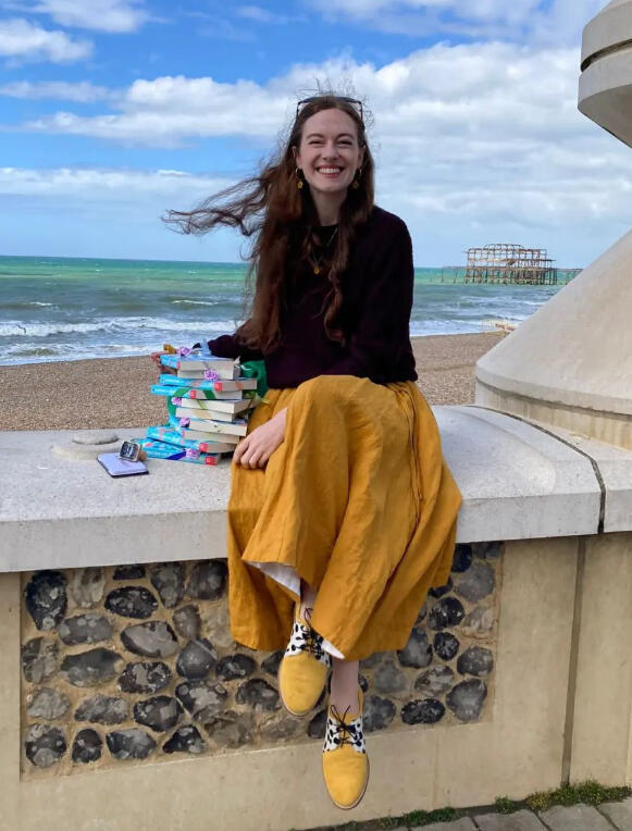 A photograph of a windswept Fiona perching on a wall with a huge stack of Looking for Emily books beside her. The sea and the old Brighton Pier are visible over her shoulder.